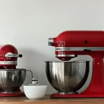 Two red stand mixers with stainless steel bowls on a wooden countertop, with white ceramic bowls on a shelf above.